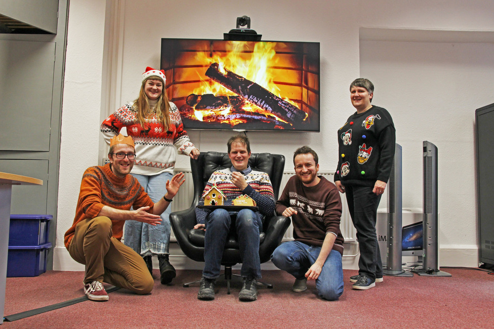 WJPS team in Christmas Jumpers stood in front of a TV Screen with a fire on